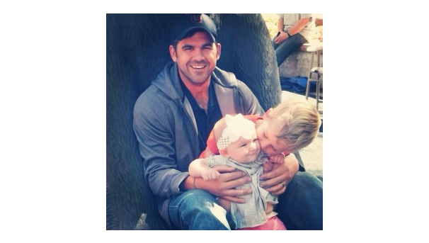 A man sitting against the base of a tree. He is holding two children: a toddler and a baby.