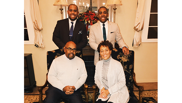 Four individuals, two sitting, two standing, smile while posing for a family photo