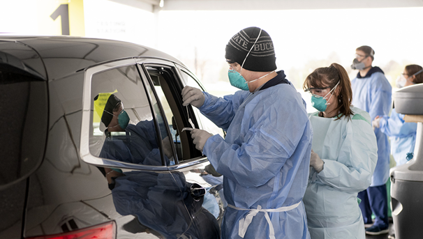 Two healthcare workers stand next to a car. One is working on administering a COVID-19 test