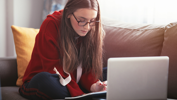 Student witting on a couch, taking notes. Her laptop sits across from her