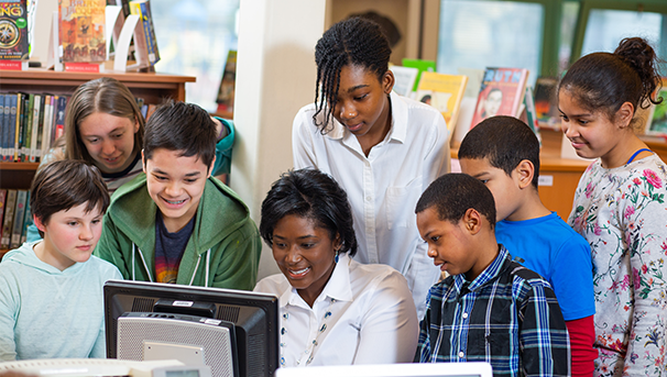Students of varying ages stand around a desktop computer at a library