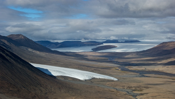 A brown landscape from the side of a mountain. There are sections of ice and snow and fluffy clouds