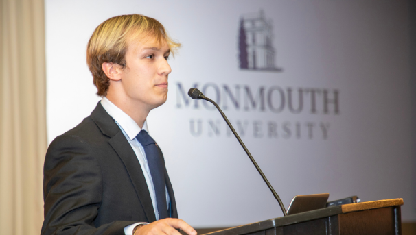 Club president presenting at a podium on campus with Monmouth University logo behind him