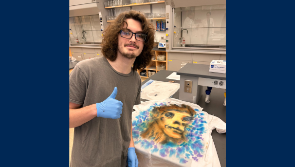 Male student giving a thumbs up next to his tie dyed lab coat that has a face on it in dye