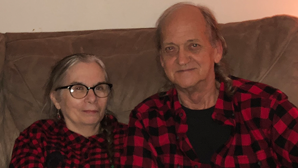 Two individuals sitting on a couch smiling for a photo wearing red black checkered shirts