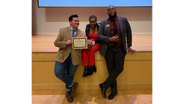 Ose and two other students at the edge of a stage. Two hold on either side of a certificate