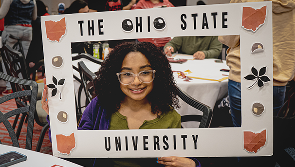 Student sitting at a table holding an Ohio State frame around their face smiling