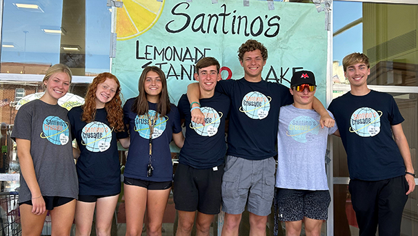 Seven individuals standing outside in front of a sign titled Santino's Lemonade Stand