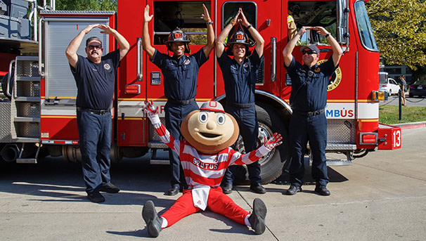 Brutus sitting on the ground with firemen doing O-H-I-O in front of a firetruck.