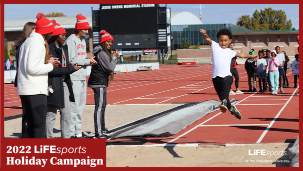 Child jumping in mid-air into the sand on the track while college students cheer him on