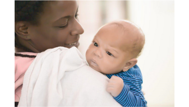A mother holds a newborn baby against her shoulder. The baby rests against a white cloth