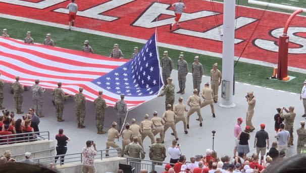 Group of uniformed service members and ROTC participants hold large American flag on football field