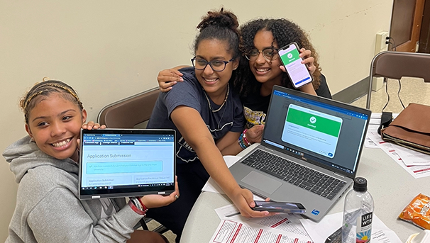 Three students holding up tablets while smiling for photo sitting at table