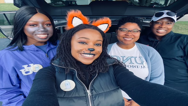 Four people posed for a selfie. They are beside an open car trunk for Trunk or Treat.