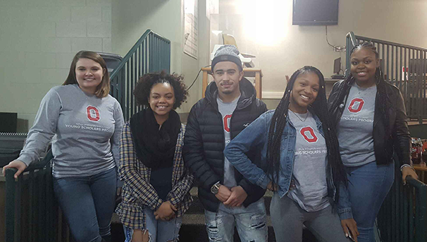 Five students standing and smiling for a group photo near a staircase