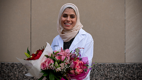 Individual wearing white coat standing outside holding bouquets of flowers