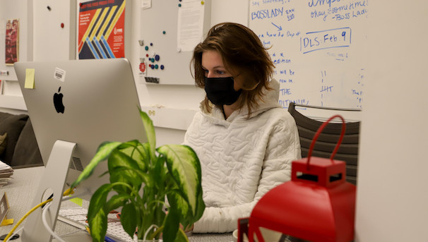 A girl wearing a mask sits at a Mac Desktop, writing. Behind her is a filled out whiteboard