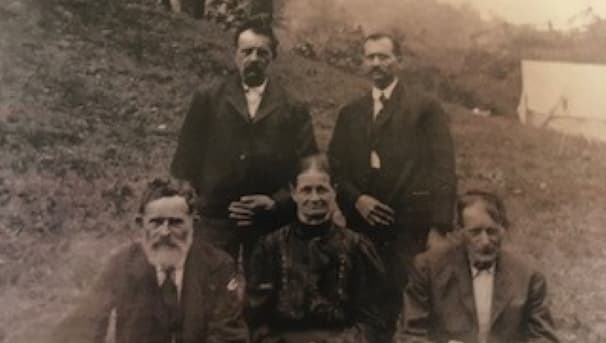 Five individuals in an black and white photo sit. They stand before a small hill and white tent