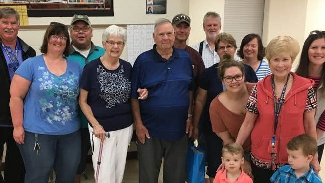 14 people gathered indoors. An older man stands in the center with the others smiling around him