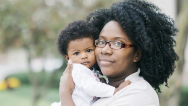 A mother with her newborn child smile towards the camera. Both wear white