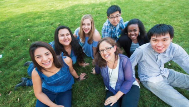 Photo of diverse group of seven students seated on grass and smiling for camera. 