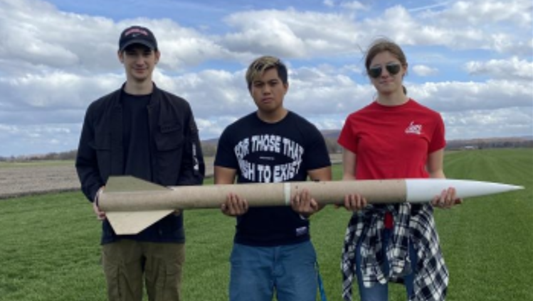 Three students holding a large model rocket in a field.