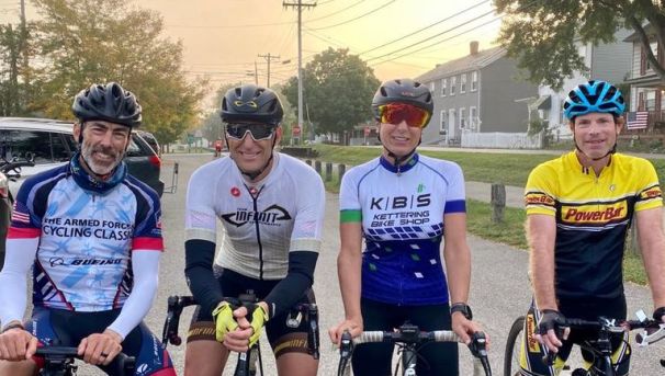 Four cyclists sit on their bikes, smiling for a photo. All wear biking gear and helmets