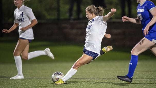 A women's soccer athlete prepares to kick the ball by being chased by her opponent.