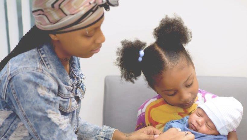 A mother helps her child hold a newborn in a hospital setting