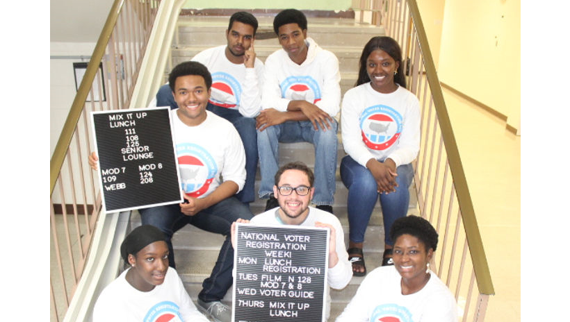 Seven students sit on a staircase. They hold two boards about voting and lunch