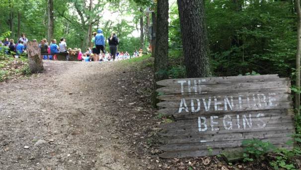 Wooden sign with 'The Adventure Begins' next to a forest path with people gathered in the background