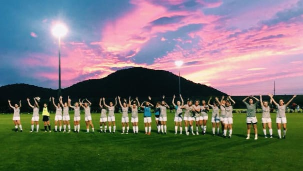 The Women's Club Soccer raise their arms towards the pink, blue, and yellow sky