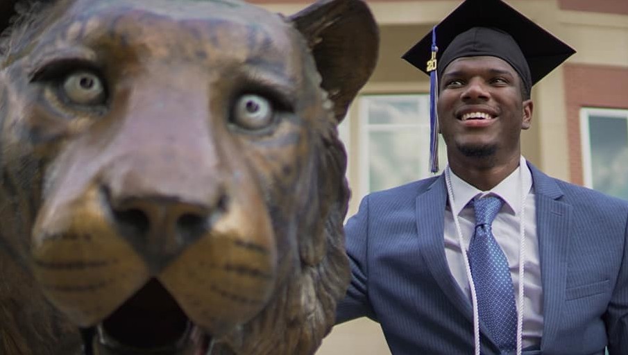 A HAAMI graduate in his cap, tassel, and honor cord smiles beside a bronze tiger statue.