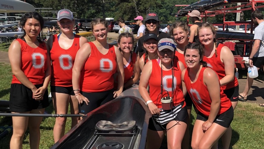 The Nov women pose together by a canoe, all wearing red tank tops with Block Os