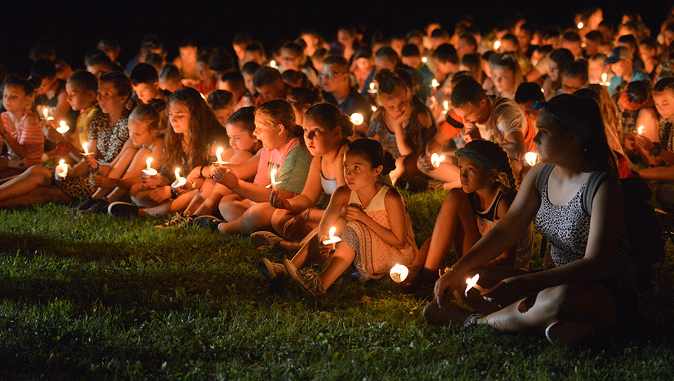 A large group of young campers hold lit candles. Outside, it is dark
