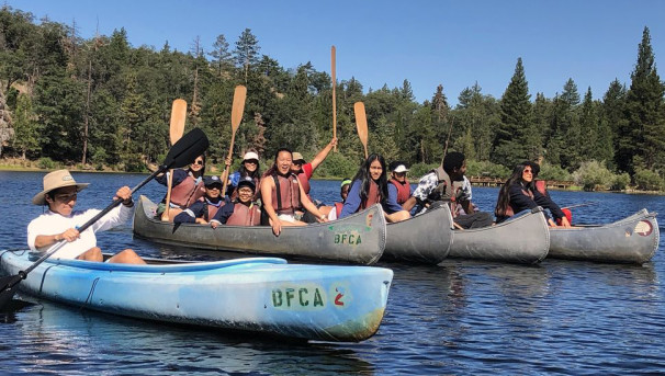 A group of students in five kayaks, waving their paddles in the air and smiling.