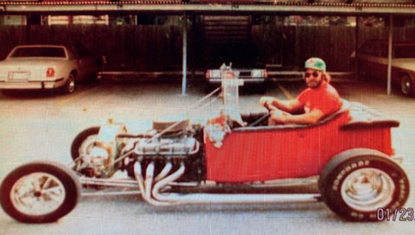 An individual sits in an old, open red car in a parking lot