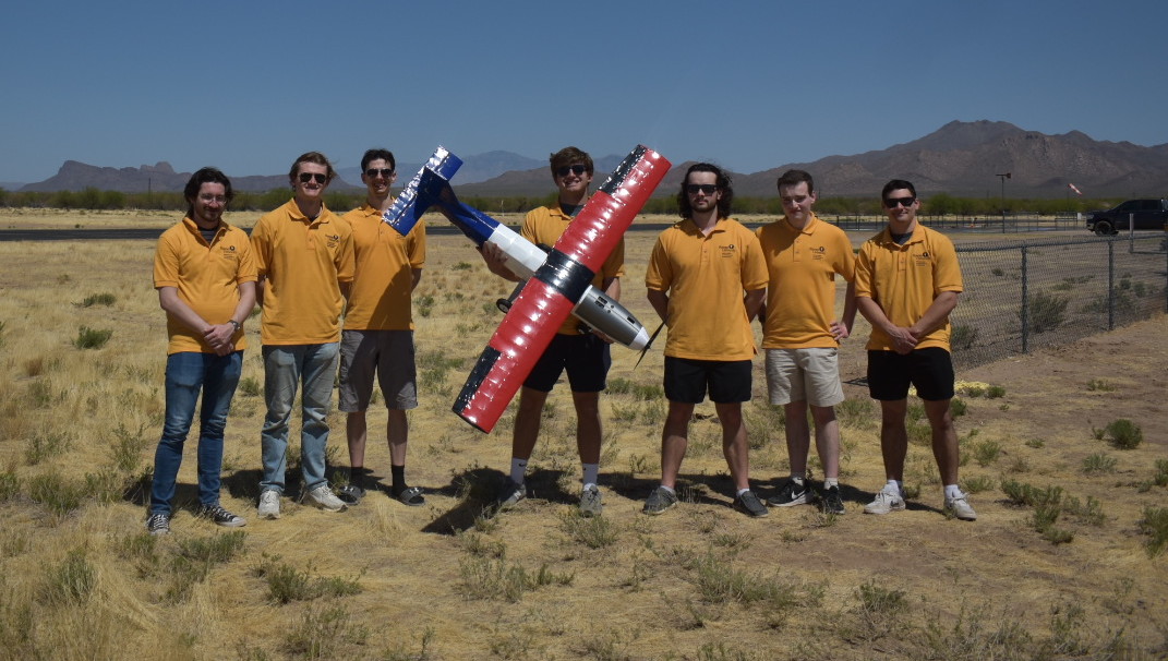 Rowan AIAA team in matcing polos in a large field holding up the plane.