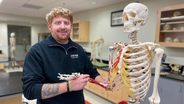 Student standing in front of a skeleton holding up its arm at the wrist