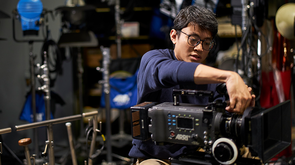 Film student adjusts a camera in a media production equipment room.