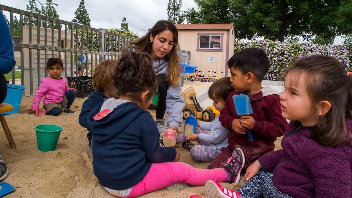 A teacher/ volunteer engaging toddlers in a sandbox activity with toys and shovels.