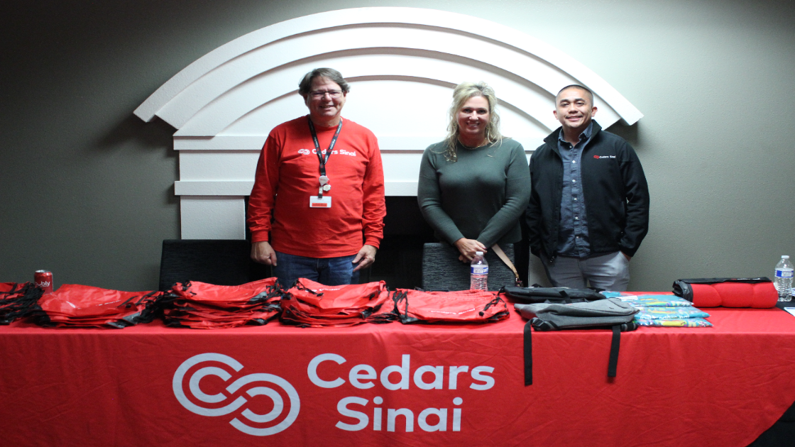Three alumni stand behind Cedars-Sinai table with red bags at alumni event.