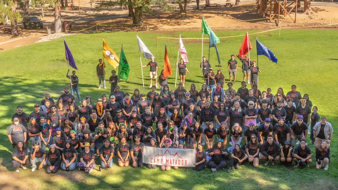 Camp Matador participants pose outdoors with flags and banner during team-building retreat.