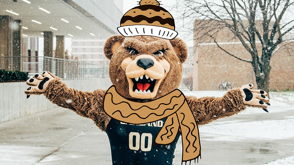 Grizz, with a cartoon hat and scarf, plays in the snow near the Elliott Tower