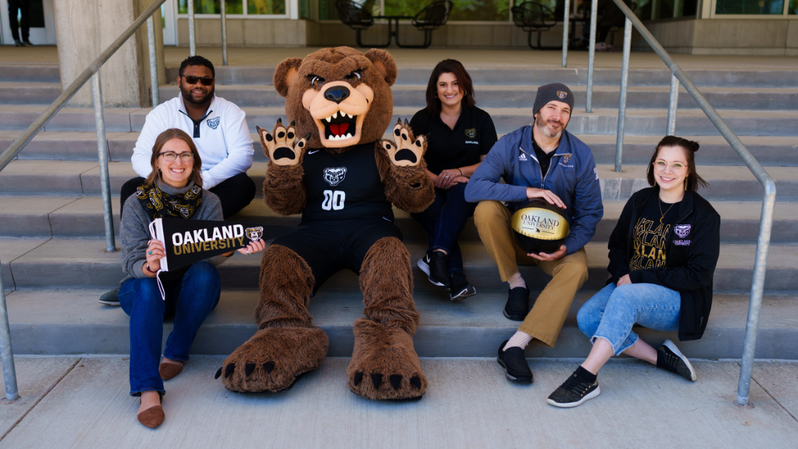 Individuals sit on the steps of the Oakland Center surrounding Grizz.