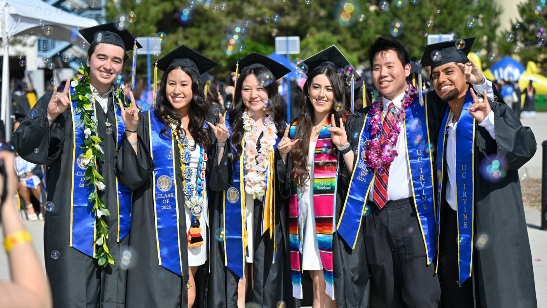 Grads pose in full regalia holding the "zot" hand sign with bubbles falling around them