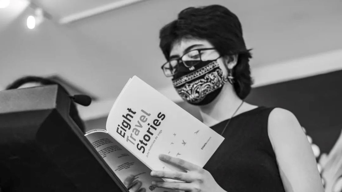 A woman standing at a poem reading a book titled Eight Travel Stories