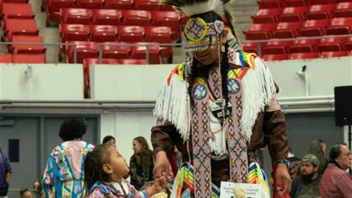 An elder and small child both in regalia hold hands and look at eachother.