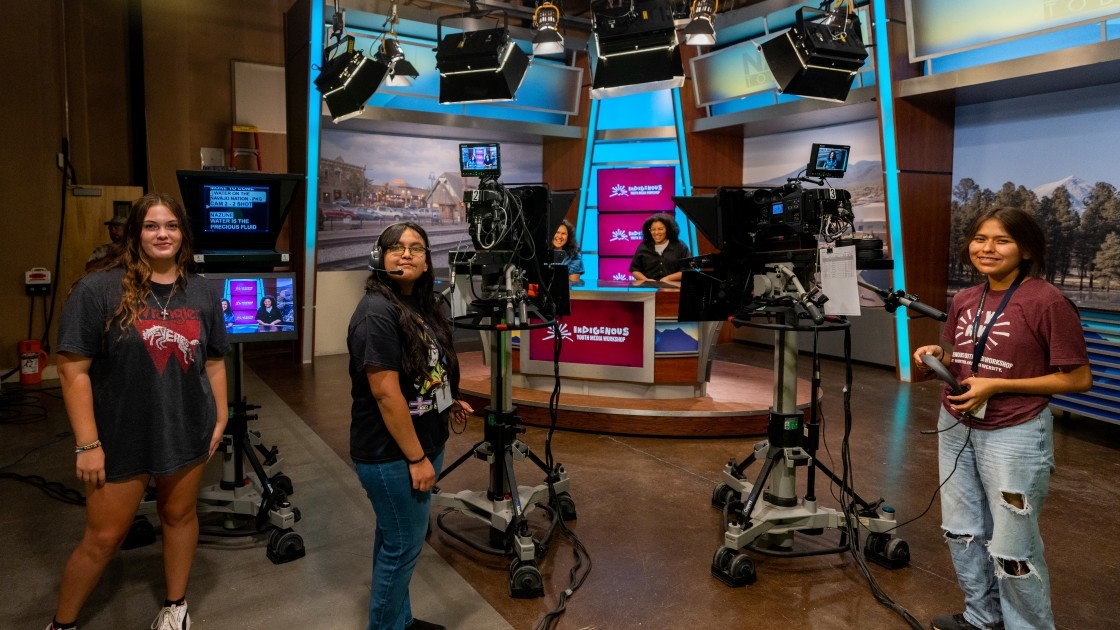 Five people in a TV studio with cameras and city skyline screens.