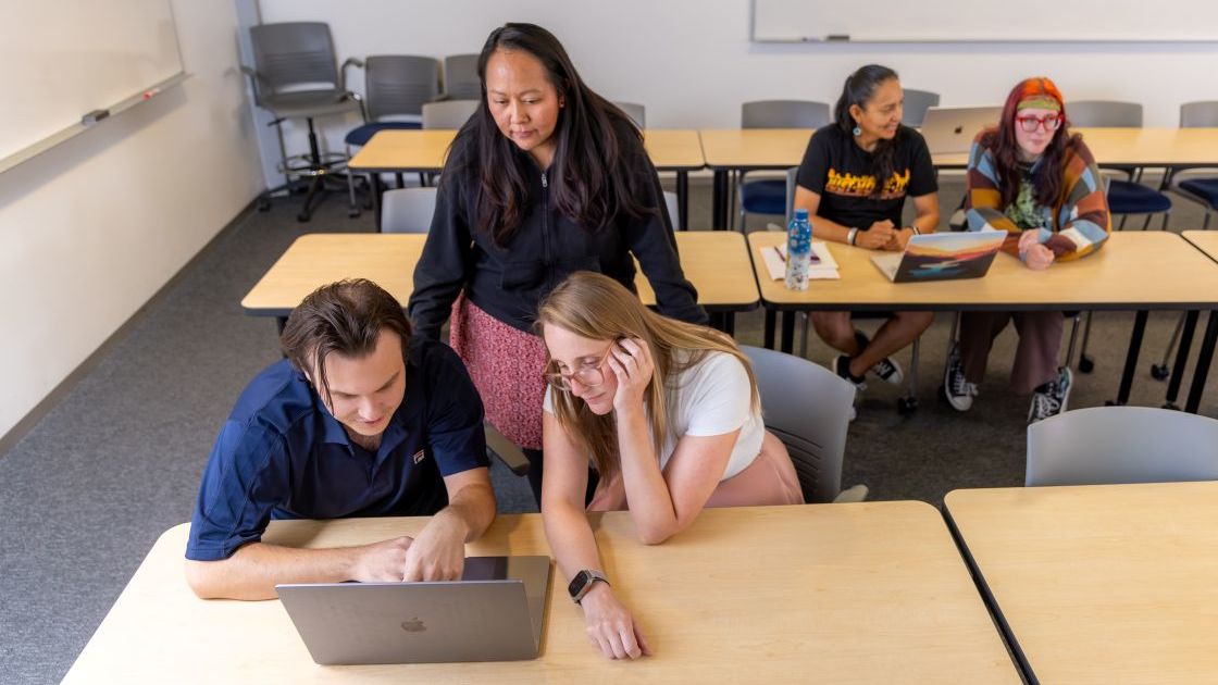 An instructor leaning over students to assist them with a coding project inside an NAU classroom.